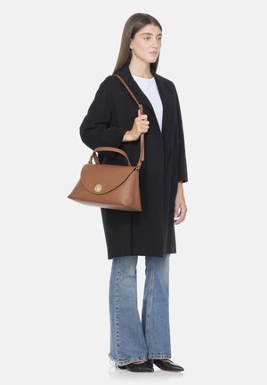 Femme portant un manteau noir et un jean évasé, tenant un sac à main en cuir marron avec une bandoulière et une anse, sur fond blanc.