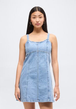 Ung kvinde med langt mørkt hår, iført guldringearmbånd og en ærmeløs lyseblå denimkjole mod en ensfarvet lys baggrund.