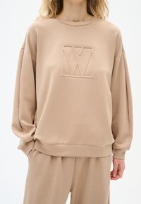 Beige sweatshirt med en texturerad logotyp på bröstet, rundad halsringning och avslappnad passform. Mjuk material, neddragna axlar och ribbade ärmslut.