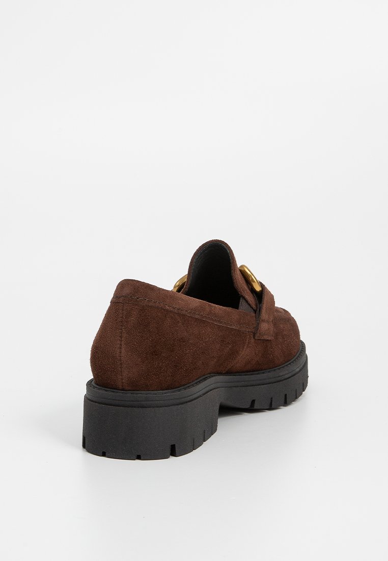 Bruine suède instapper met een dikke zwarte rubberen zool, voorzien van een decoratieve strap en accenten van goudkleurig metaal. De achterkant benadrukt de soepele textuur.
