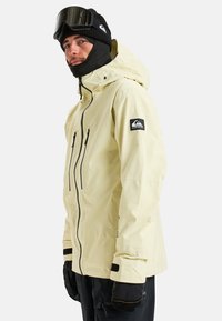 Helle gelbe wasserdichte Jacke mit Reißverschlusstaschen, verstellbaren Manschetten und einer Kapuze, kombiniert mit schwarzen Handschuhen und einer Skimaske.