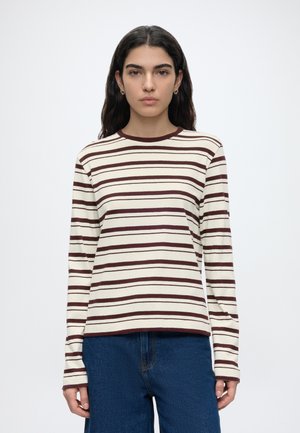 Femme aux longs cheveux foncés portant des boucles d'oreilles créoles dorées, une chemise blanche à manches longues avec des rayures horizontales marron et un jean bleu, debout.
