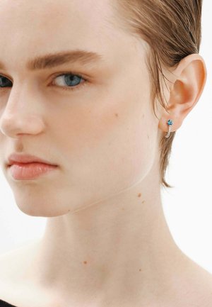 Pendiente de aro de plata con un acento de gema azul, que presenta una textura suave y un diseño sencillo. Primer plano de la oreja y la línea de la mandíbula de un modelo.