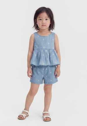 Μικρό παιδί όρθιο, φοράει μπλε αμάνικη μπλούζα και αντίστοιχα σορτς με λευκά ανθάκια και λευκά σανδάλια.