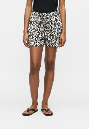 Mujer con pantalones cortos de cintura atada con estampado en blanco y negro, top sin mangas negro y sandalias negras planas, de pie sobre un fondo liso.