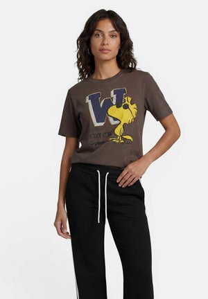 Frau trägt ein braunes T-Shirt mit einem gelben Cartoon-Vogel, schwarze Sonnenbrille und den Text "Stay Cool," kombiniert mit schwarzen Hosen mit Kordelzug.