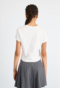 T-shirt en coton blanc à manches courtes, associé à une jupe plissée grise. La vue de dos montre un design simple et un tissu léger.
