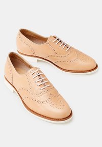 Beige nahka brogue -kengät, joissa on reikäkoristeita, pyöreä kärki, valkoiset nauhat ja kontrastivalkoinen pohja.