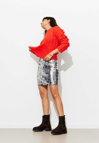 Pull rouge en maille torsadée, jupe argentée à paillettes et bottines noires à semelles épaisses. Le mannequin pose devant un mur blanc.