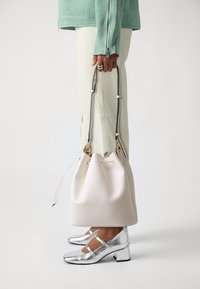 Borsa a secchiello bianca in pelle con chiusura a coulisse e dettagli dorati, tenuta da una persona che indossa una giacca menta e scarpe con tacco argentate.