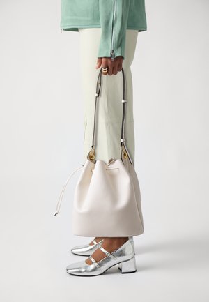 Bela usnjena torba v obliki vedra z zategovanjem na vrhu in zlatimi detajli, ki jo drži oseba v mentol zeleni jakni in s srebrnimi čevlji na visokem petku.