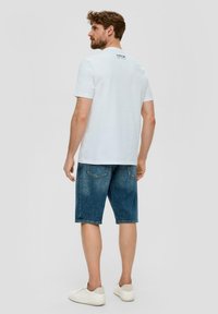 Camiseta blanca de manga corta con logo en la espalda, combinada con pantalones cortos de denim azul y zapatillas blancas, de pie contra un fondo neutral.