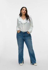 Blouse blanche avec bordures à volants, manches longues et fermeture à boutons ; portée avec un jean en denim bleu taille haute légèrement évasé au bas.