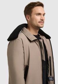 Veste matelassée beige avec un col en polaire noir, fermeture éclair à l'avant et poches latérales, avec un logo de la marque sur la poitrine.