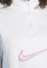Pull-over demi-zippé Nike en blanc et bleu clair, avec un logo Swoosh rose brodé. Tissu doux avec une texture lisse.