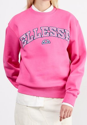 Woman wearing bright pink sweatshirt with white "ELLESSE" logo across chest and white shirt layered underneath, paired with light-colored pants.