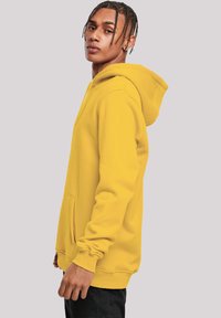 Yellow hoodie made of soft fabric, featuring a drawstring hood, kangaroo pocket, and ribbed cuffs. Model stands in profile against a plain background.