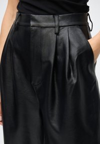 Schwarze Lederhose mit hoher Taille, Falten und Seitentaschen. Die Struktur ist glatt mit einem leichten Glanz.