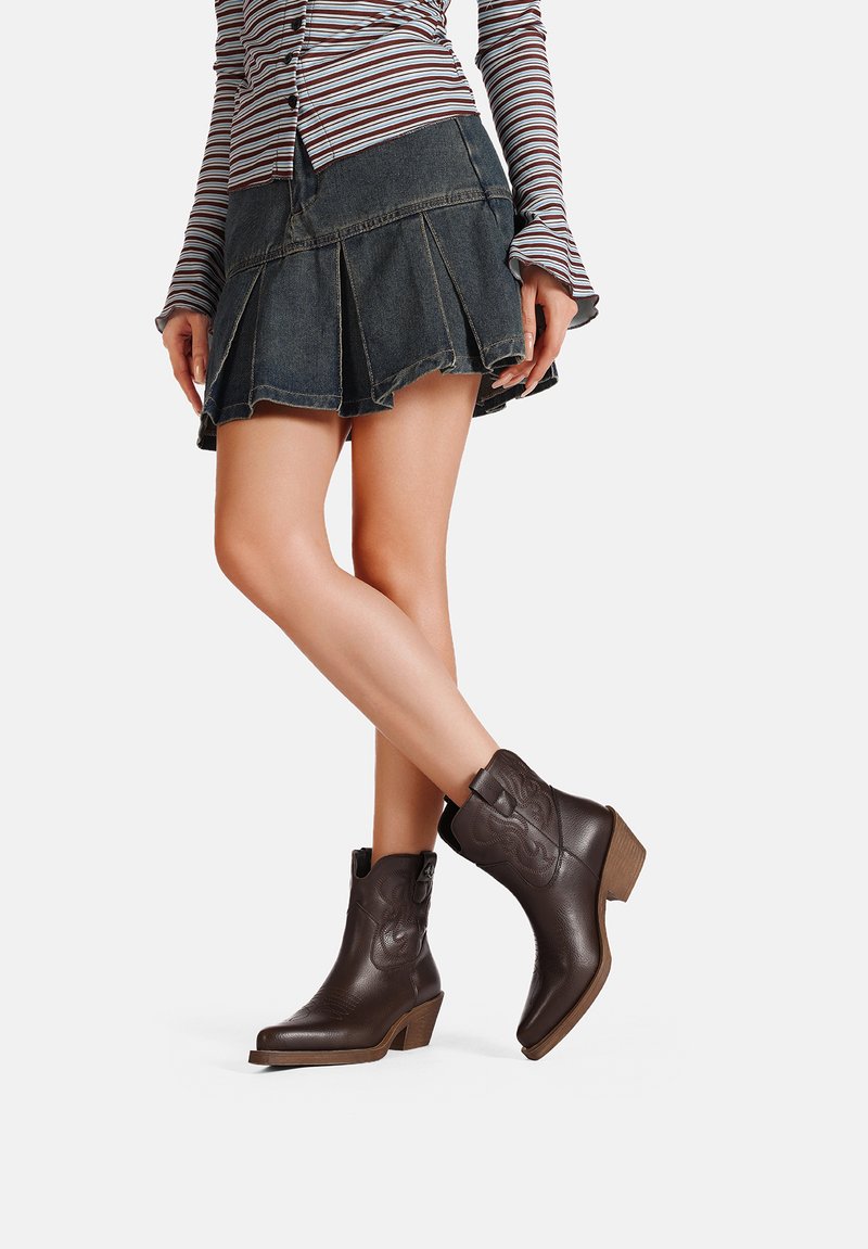 Stivaletti in pelle marrone con cuciture decorative, punta rotonda e tacco impilato. Abbinati a una gonna plissettata in denim.