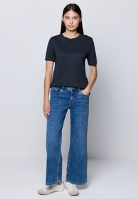 Giovane donna in piedi con la mano in tasca, che indossa una maglietta nera, jeans larghi blu e sneakers bianche su uno sfondo neutro.