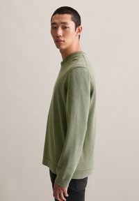 Oliwkowy sweter z długim rękawem i okrągłym dekoltem, z prążkowanymi mankietami i dołem, wykonany z teksturowanej dzianiny. Noszony z czarnymi spodniami.