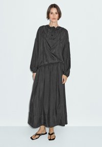 Robe longue gris foncé avec une coupe décontractée et un col à cordon ; associée à de simples sandales noires. Tissu lisse, accents minimes, silhouette élégante.