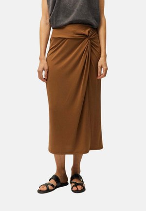 Mujer con una falda midi marrón con detalle de nudo lateral, top gris sin mangas y sandalias planas negras con tiras cruzadas.