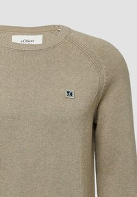 Beżowy sweter z dzianiny z okrągłym dekoltem i małą kwadratową metką z logo s.Oliver po lewej stronie klatki piersiowej.