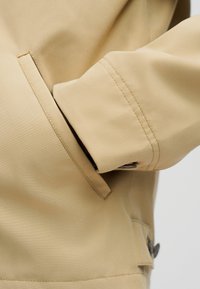 Manche de veste beige avec coutures visibles et détail de bouton, main partiellement insérée dans une poche latérale fendue.
