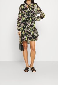 Sort blomsterkjole med lange ærmer, bølget kant og bindebånd ved halsen. Udsmykket med lilla og grønne blomster. Bæres med en sort quilted handbag og sandaler.