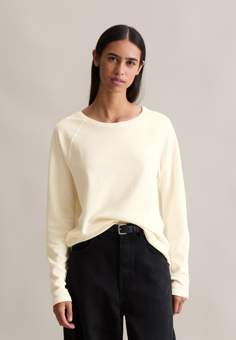 Cream-colored long-sleeve top with a round neckline and raglan sleeves, paired with high-waisted black jeans and a black belt.