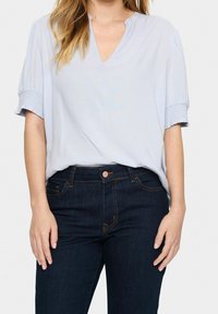 Blusa azzurro chiaro con scollatura a V e maniche corte arricciate. Abbinata a jeans in denim scuri con tasca frontale e bottone.