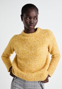 Pull jaune duveteux avec un col rond et des manches longues, présentant un tissu texturé et doux. Porté avec un pantalon à carreaux gris.