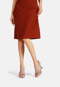 Sandali con tacco alto beige con cinturini intrecciati a incrocio e design con punta aperta, abbinati a un vestito color ruggine di lunghezza al ginocchio. Tessuto liscio.