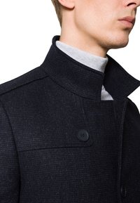 Czarny wełniany płaszcz z wysokim kołnierzem, teksturowanym materiałem i zapięciem na jeden guzik. Widoczne szwy i slim-fit design.