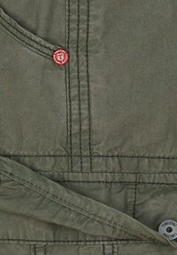 Overoles de algodón verdes con costuras en contraste. Botón redondo rojo destacado y pequeña etiqueta circular en la parte inferior derecha. Textura suave.