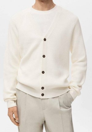 Homme portant un cardigan côtelé de couleur crème avec cinq boutons foncés sur une chemise blanche et un pantalon beige clair, main dans la poche.