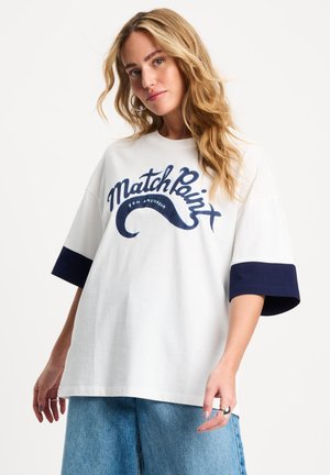 Blonde vrouw draagt een wit oversized T-shirt met navy manchetten en de tekst "Match Point", gecombineerd met blauwe spijkerbroeken.