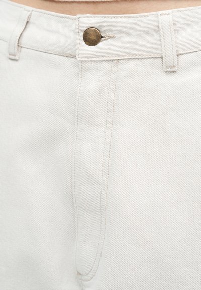 Εγγύς πλάνο της μέσης λευκών τζιν από τζιν με ορειχάλκινο κουμπί και ορατές λεπτομέρειες ραφής.