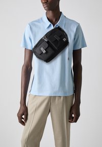 Borsa a tracolla nera in nylon con molteplici tasche, chiusura con zip e dettagli grigi, indossata sopra una camicia azzurro chiaro con colletto e pantaloni beigi.