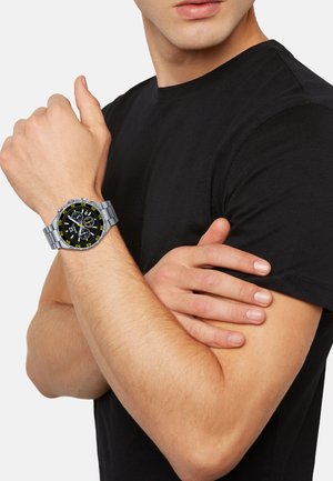 Sølv rustfrit stål ur med en sort og gul urskive, der har chronograph funktioner, båret på et håndled mod en sort t-shirt baggrund.