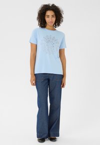 T-shirt bleu clair à manches courtes avec un motif de découpes circulaires au centre, associé à un jean en denim bleu ample et des chaussures noires.