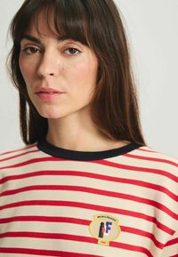 T-shirt rayé avec des bandes horizontales rouge et crème, un col noir, et un patch logo avec une figure stylisée et des détails textuels.