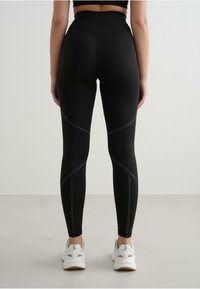 Colanți negri cu talie înaltă, dintr-un material elastic, cu linii elegante și un design texturat, asortați cu adidași albi. 