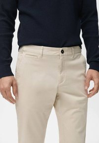 Beige chinos with a slim fit, featuring a flat front, side pockets, and a single button closure. Paired with a textured navy sweater.