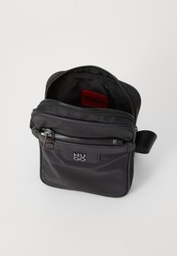 Sort nylon crossover taske med lynlås, der har en struktureret ydre og indre foring, samt et rødt HUGO logo mærke. Justerbar rem.