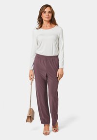 Licht lange mouwen shirt en donkere pruimen kleur elastische taillebroek. Model houdt een kleine beige handtas vast. Glad stof met een ontspannen pasvorm.