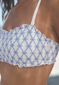 Witte bralette top met blauwe bloemenborduurwerk, gegolfde randen en dunne schouderbandjes. Close-up van de stof en ontwerpdetails.