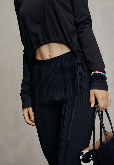 Top preto cropped de manga longa com laterais franzidas, combinado com leggings pretas de cintura alta em malha texturizada. Pulseira colorida de contas e bolsa.