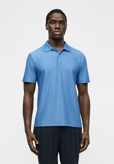 Άνδρας που στέκεται προς τα εμπρός φορώντας ένα ανοιχτό μπλε πλεκτό πόλο και σκούρο παντελόνι, με φόντο ένα απλό λευκό υπόβαθρο.